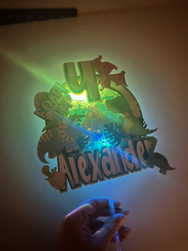 Custom dinosaur "Roar Im 4" cake topper with greens and blue dinosaurs and colors with a shaker and lights