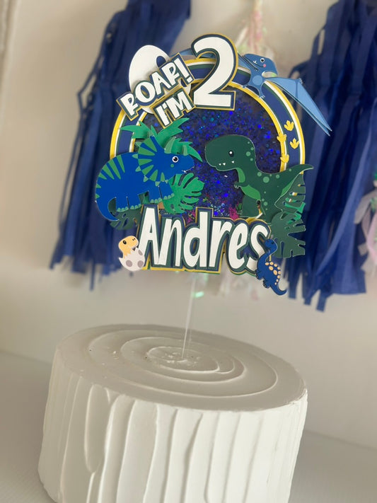 Decorative cake topper with dinosaurs and text on a white stand against a neutral background